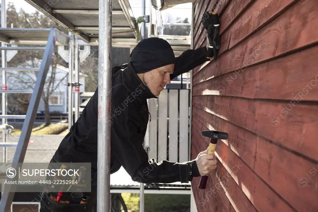 Side view of confident repairman hammering wooden wall at construction site