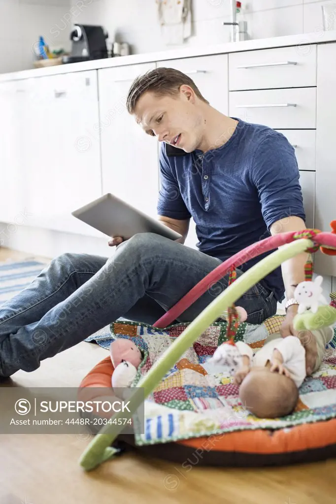 Father using digital tablet and mobile phone while baby sleeping on mat in kitchen