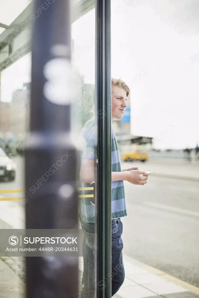 Young man holding mobile phone while standing at bus stop