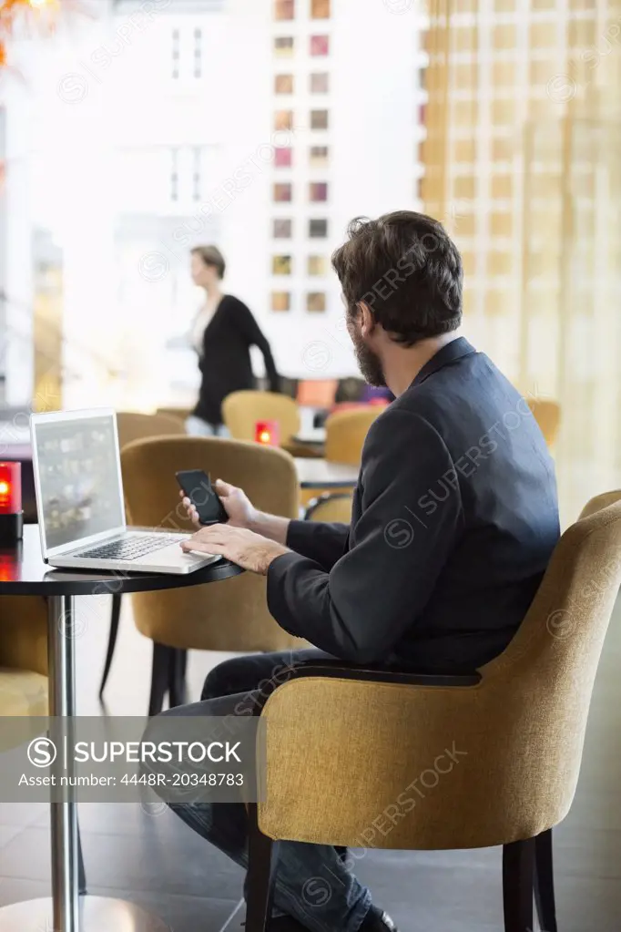 Side view of businessman with mobile phone and laptop sitting in hotel restaurant