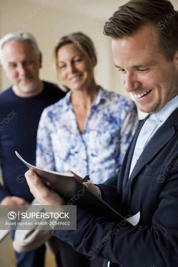 Smiling male real estate agent writing on document with couple standing in background
