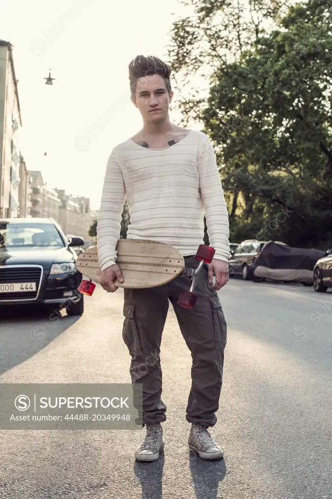 Full length portrait confident male high school student with skateboard standing on city street
