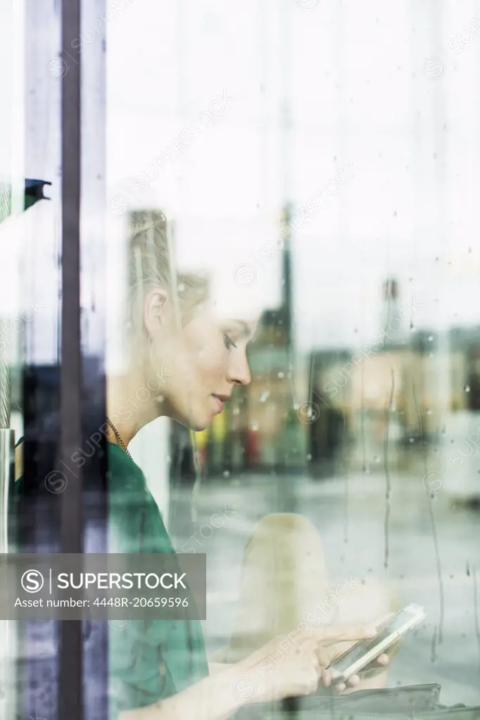 Young using smart phone while sitting on sill seen through glass at railroad station