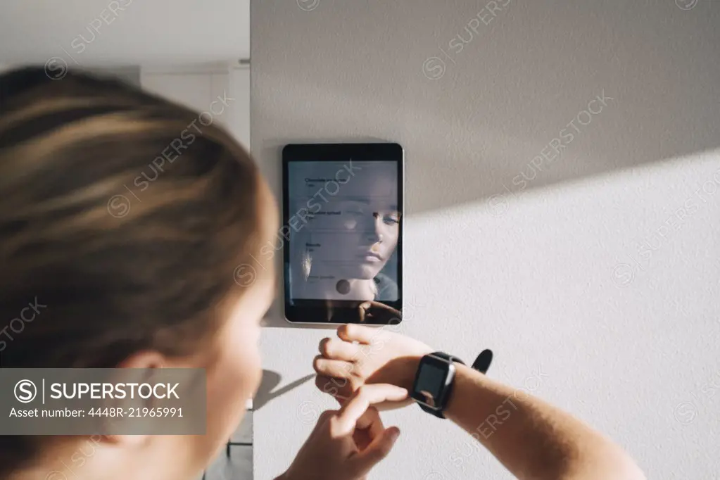 Teenage girl adjusting smart watch with digital tablet mounted on white wall at home