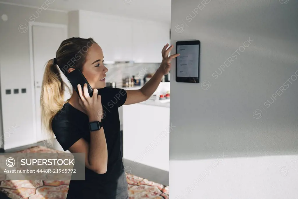 Teenage girl talking on smart phone while using digital tablet mounted on white wall at home