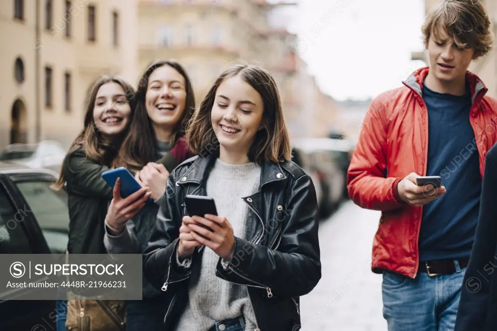 Smiling friends using mobile phones while walking on sidewalk in city