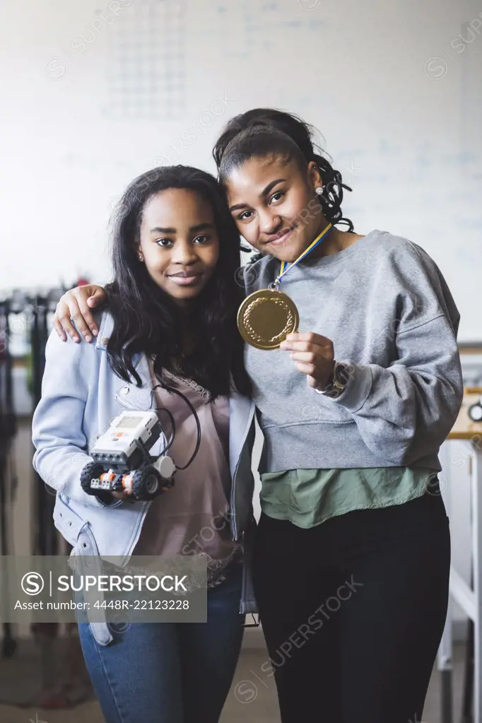 Portrait of successful female students holding robot and gold medal while standing in classroom at high school