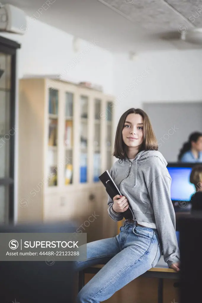 Portrait of confident female student sitting with books on desk in classroom at high school