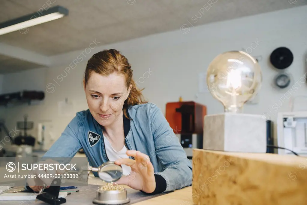 Confident redhead female engineer looking through magnifying glass at workbench in creative office