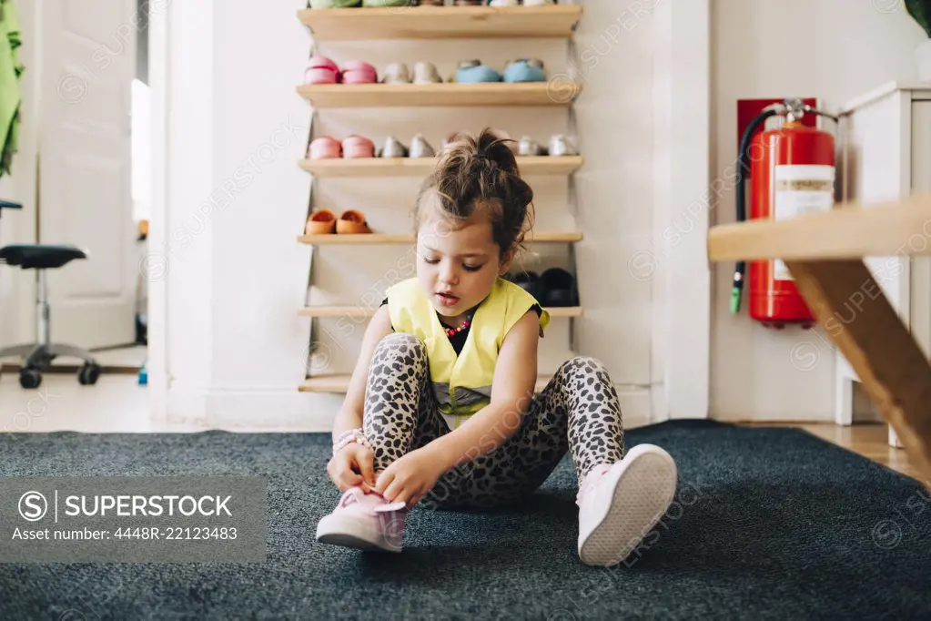 Girl sitting on carpet wearing shoes against rack in cloakroom at child care
