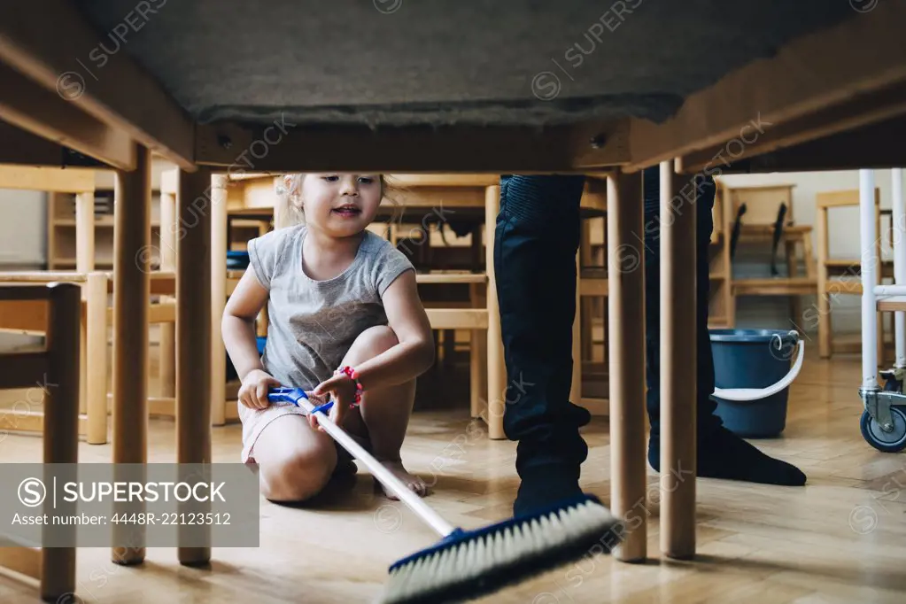 Full length of girl cleaning hardwood floor below table by teacher standing in classroom