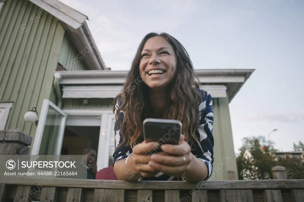 Happy mid adult woman using mobile phone while leaning on railing at porch