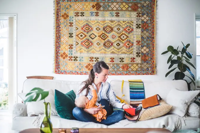 Fashion designer breastfeeding daughter while looking at fabric swatch on sofa in living room