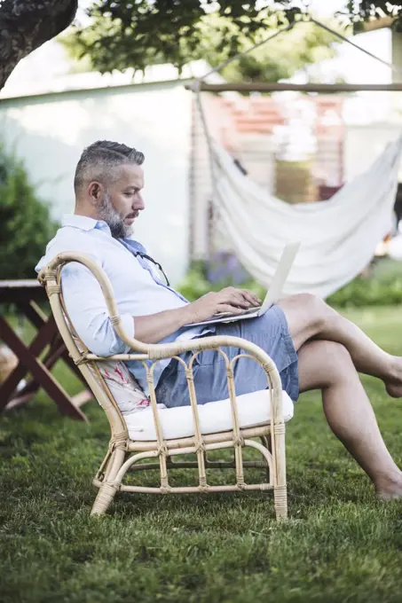 Side view of mature man working on laptop while sitting on chair in backyard