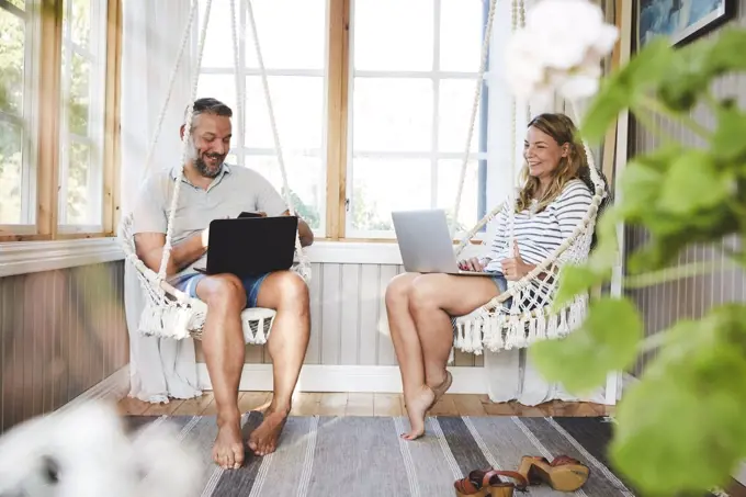 Smiling couple using laptops while sitting on rope swing in log cabin