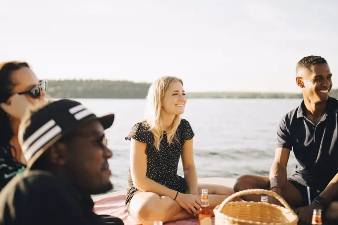 Cheerful multi-ethnic friends talking while sitting on jetty at lake in summer