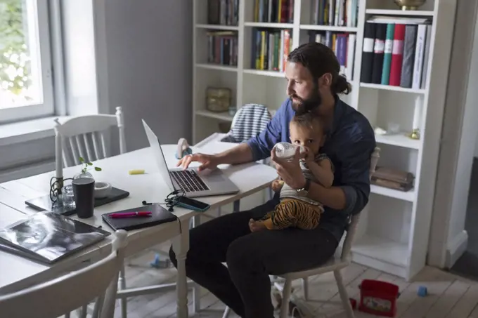 Father feeding baby boy while using laptop at home