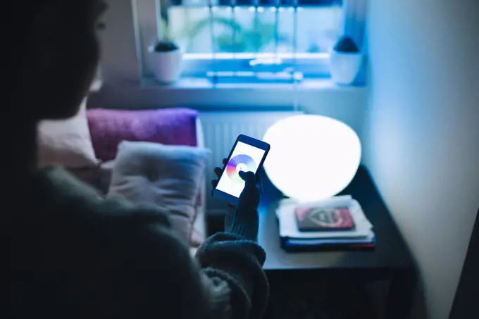 Cropped image of teenage girl using smart phone while standing by illuminated bed in room