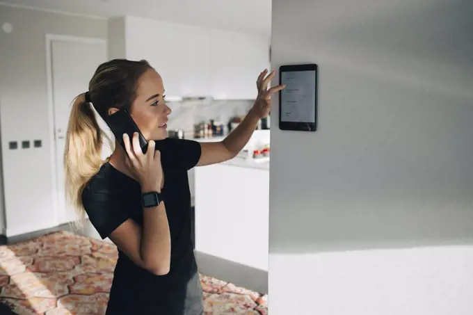 Teenage girl talking on smart phone while using digital tablet mounted on white wall at home