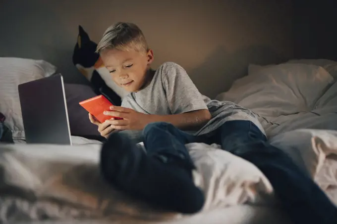 Full length of boy using mobile phone reclining with laptop on bed at home
