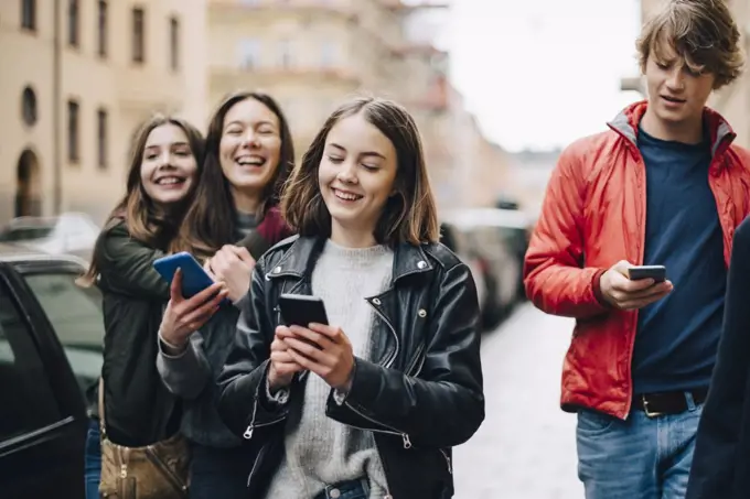 Smiling friends using mobile phones while walking on sidewalk in city