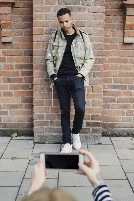 Cropped hands of young woman photographing fashionable man standing with hands in pockets against brick wall