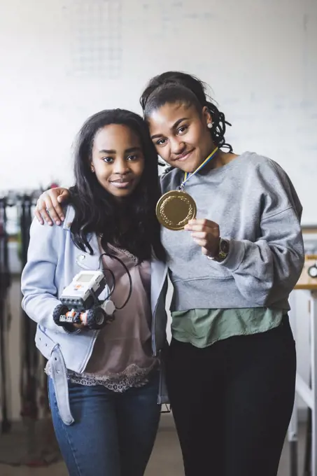 Portrait of successful female students holding robot and gold medal while standing in classroom at high school