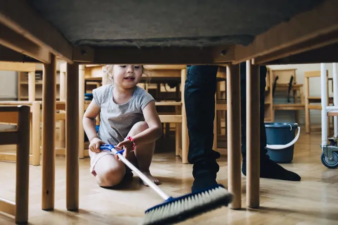 Full length of girl cleaning hardwood floor below table by teacher standing in classroom
