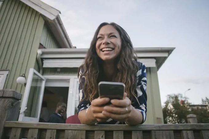 Happy mid adult woman using mobile phone while leaning on railing at porch