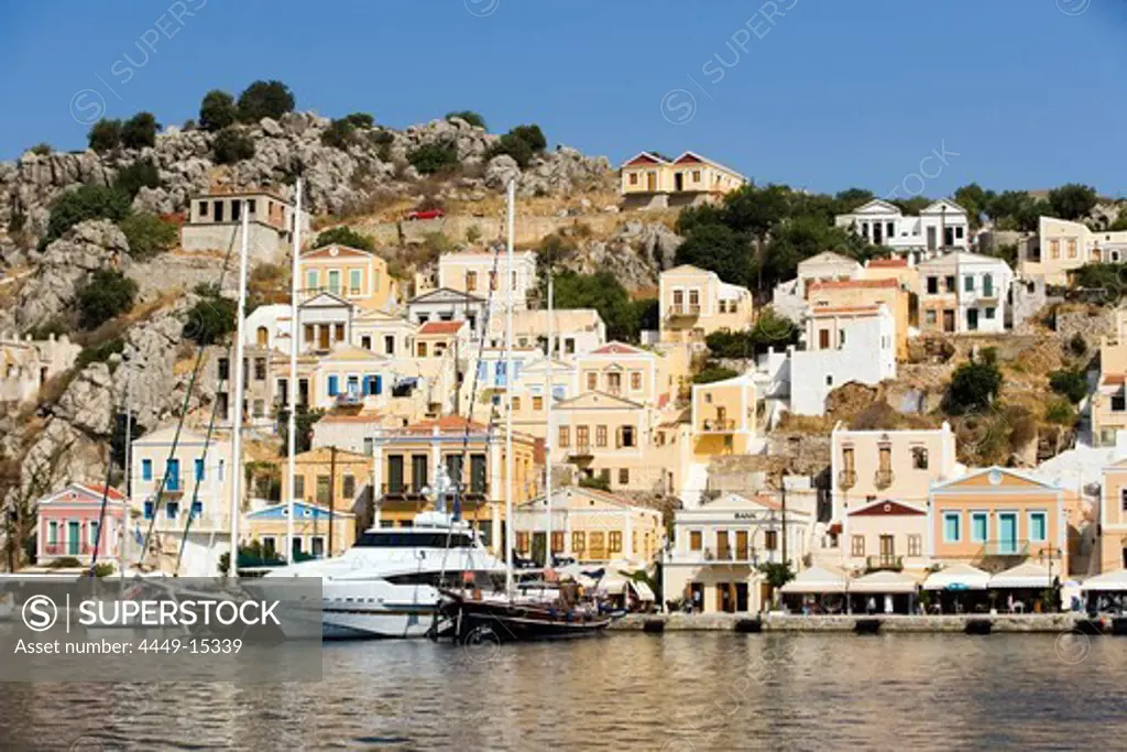 Yacht and sailing boats anchoring at quay of harbour Gialos, picturesque mansions at mountainside in background, Simi, Symi Island, Greece