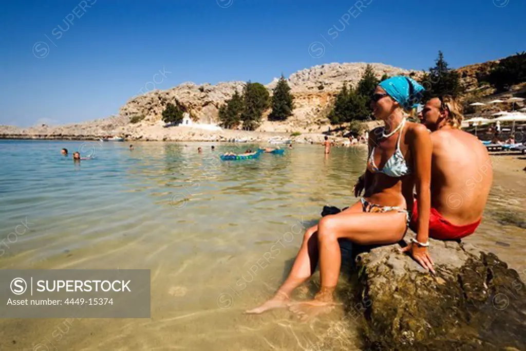 Couple sitting on rocks and talking, Saint Paul's Bay (Agios Pavlos), Lindos, Rhodes, Greece
