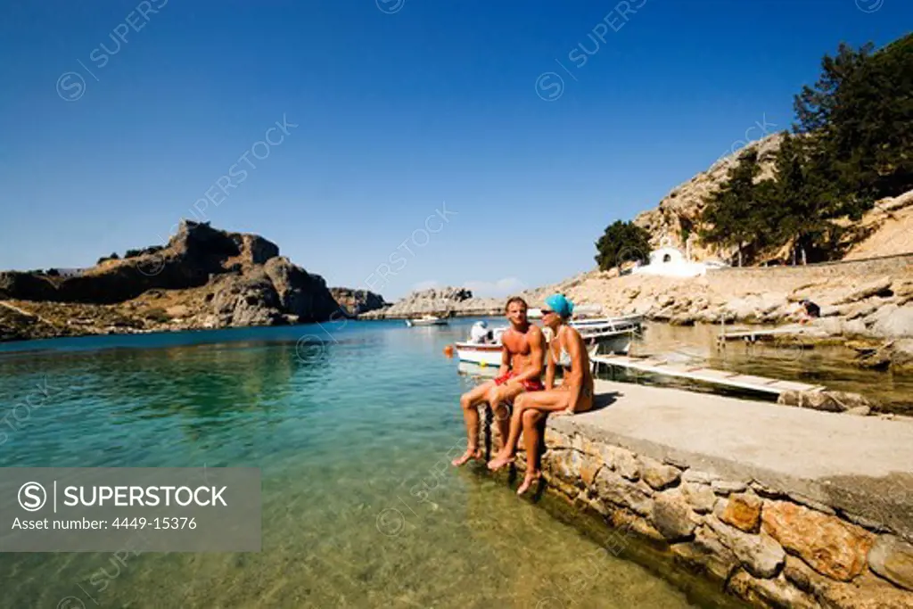 Couple sitting on rocks and talking, Saint Paul's Bay (Agios Pavlos), Acropolis in the back, Lindos, Rhodes, Greece