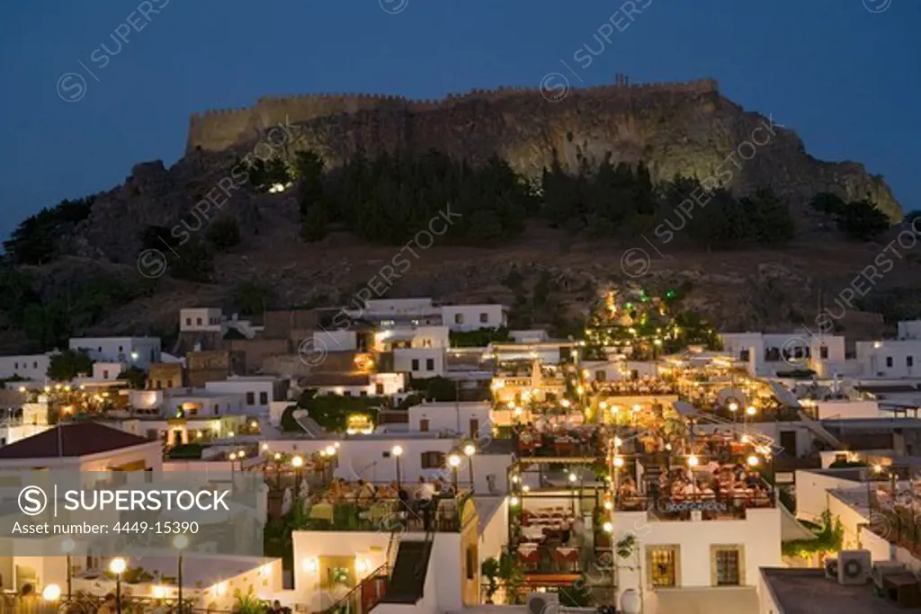 View over illuminated town in the evening to Acropolis, people sitting on terraces of restaurants, Lindos, Rhodes, Greece