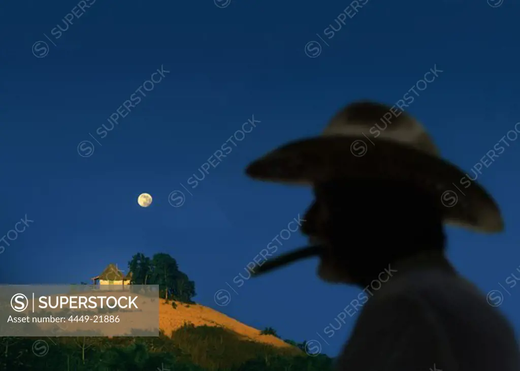 Farmer with hat smoking a cigar in the foreground, moon rising over tobacco fields, Vinales Valley, Pinar del Rio, Cuba, Carribean
