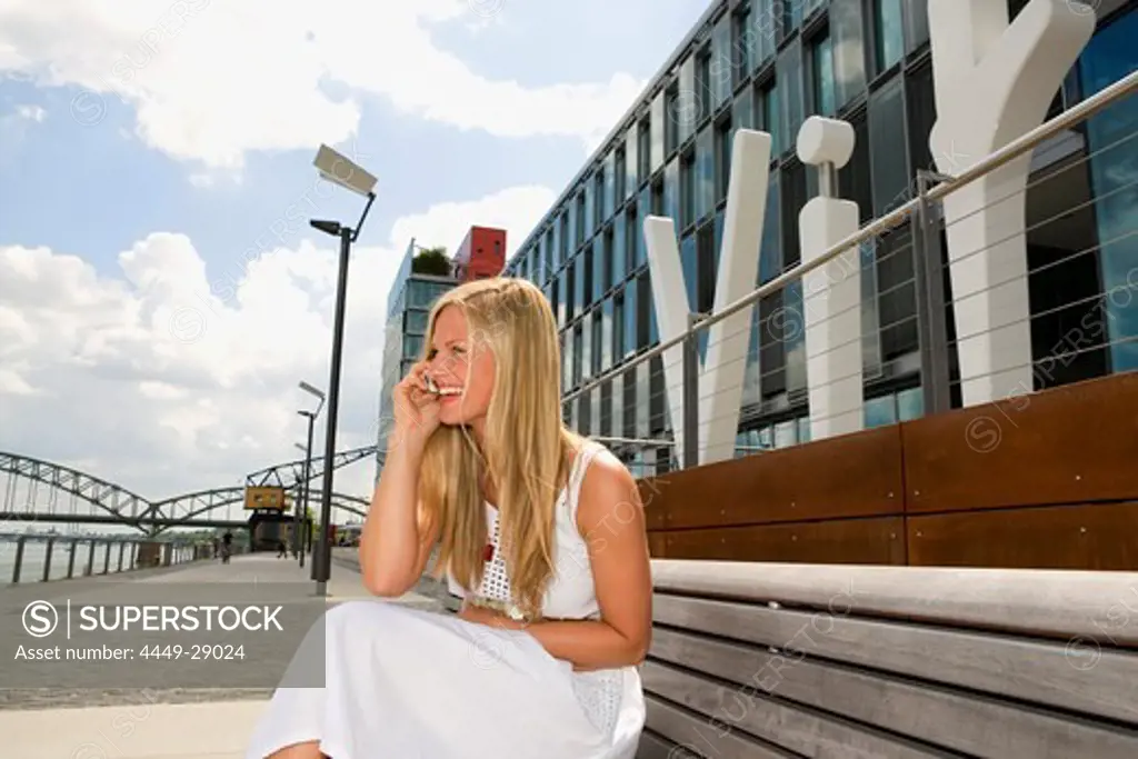 Young woman sitting on a bench while phoning with a mobile phone, Cologne, North Rhine-Westphalia, Germany