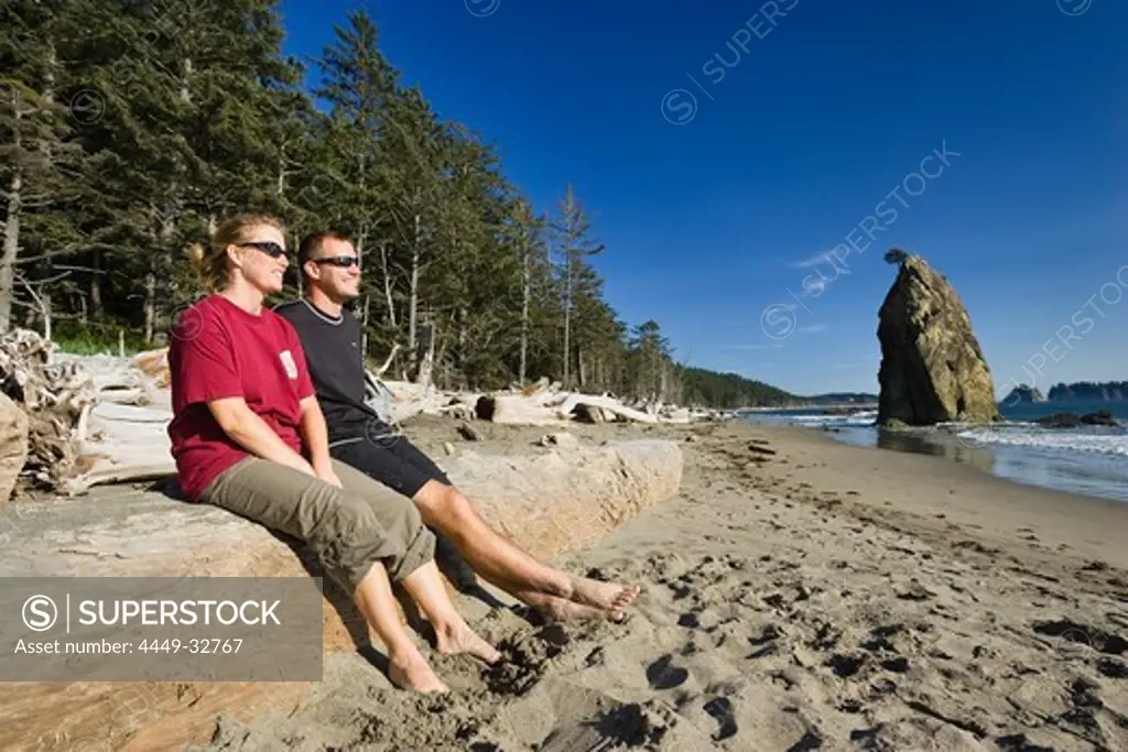 Young couple sitting on driftwood at Rialto Beach under blue sky, Olympic Peninsula, Washington, USA