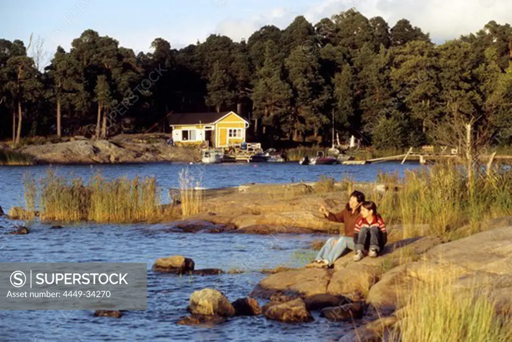 A man and a child sitting at the shore of a skerry in the light of the evening sun, South Finland, Europe