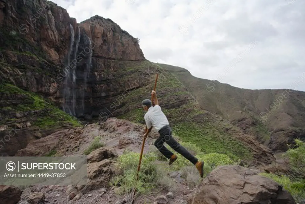 Man jumping with canarian crook in front of the waterfall Cascada el Escobar, El Risco valley, Parque Natural de Tamadaba, Gran Canaria, Canary Islands, Spain, Europe