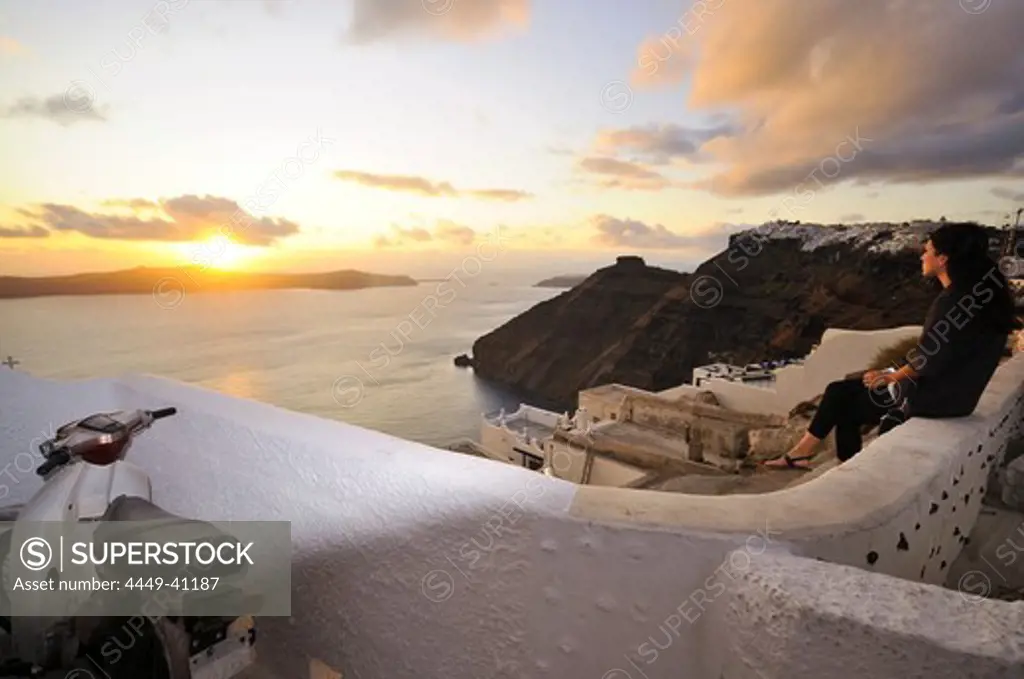 Woman sitting on a wall looking at the setting sun, Firostefani, island of Santorin, the Cyclades, Greece, Europe