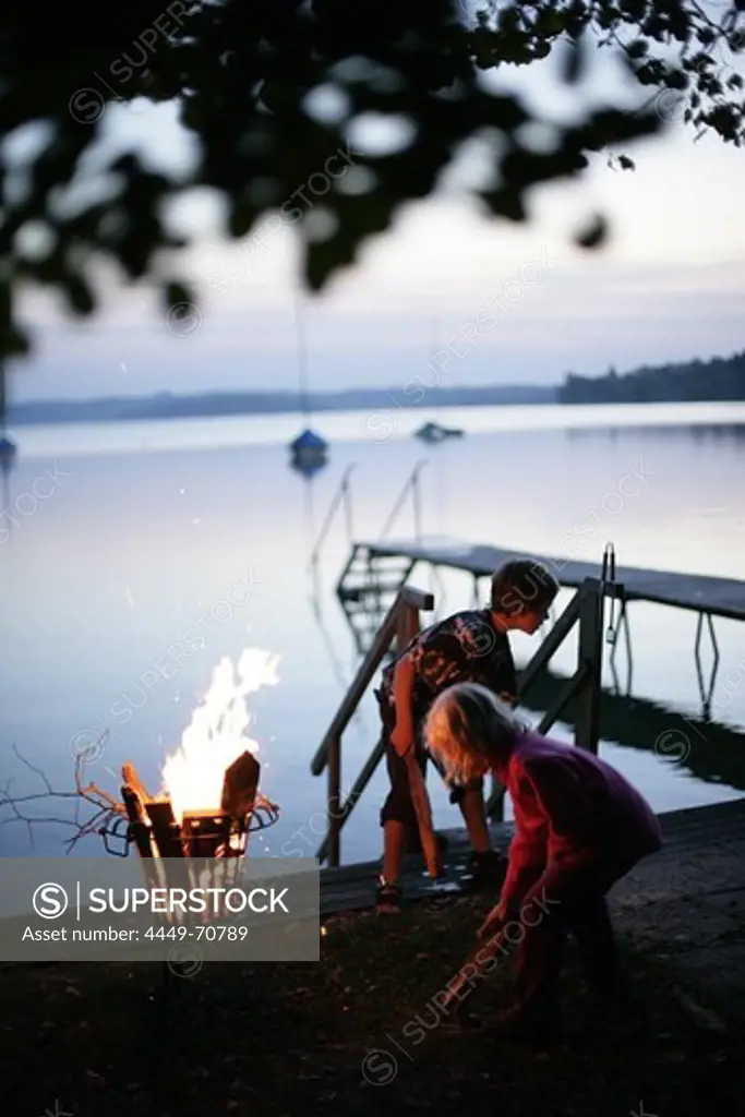 Two children standing around a fire near the jetty, evening, Lake Woerthsee, Upper Bavaria, Bavaria, Germany