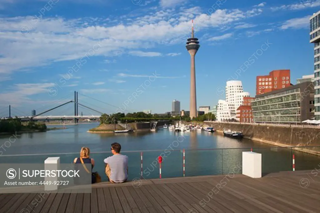 Couple sitting on a bridge at media harbour, view to Rhine tower and Neuer Zollhof, Duesseldorf, North Rhine-Westphalia, Germany, Europe
