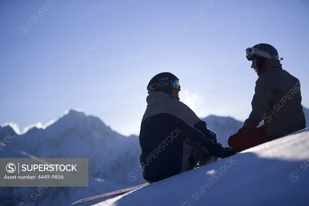 Young man and woman sitting on snow, looking mountain panorama, Kuehtai, Tyrol, Austria