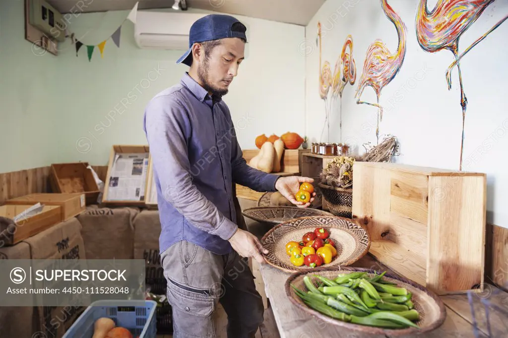Japanese man wearing cap standing in farm shop, holding bowl with fresh vegetables.