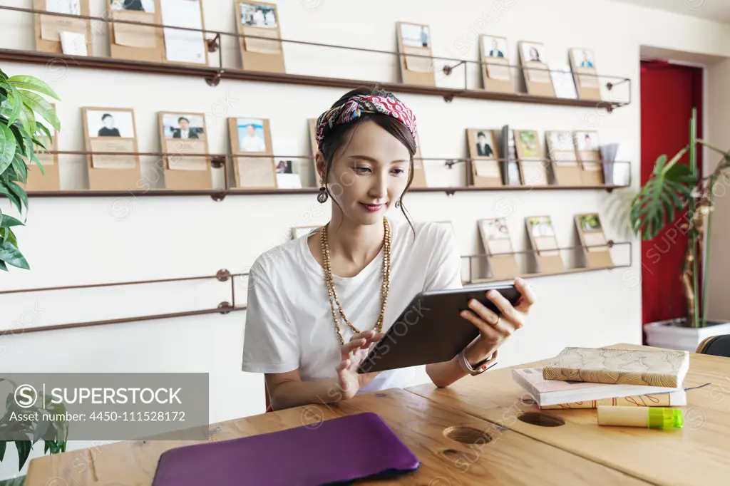 Female Japanese professional sitting at table in a co-working space, using digital tablet and laptop, holding book.