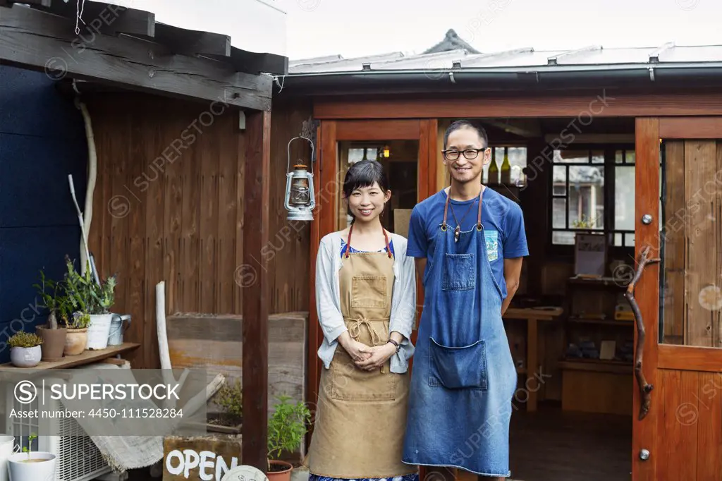 Japanese woman and man wearing blue apron standing outside a leather shop, smiling at camera.