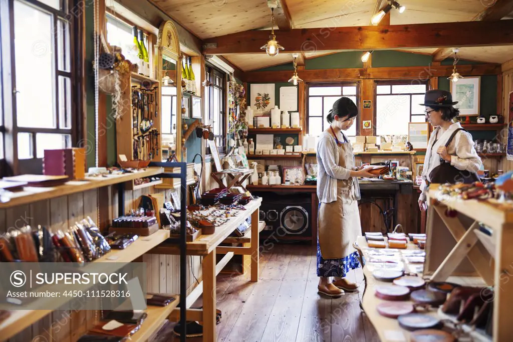 Japanese woman wearing apron standing in a leather shop, holding tan leather clutch bag, talking to customer.