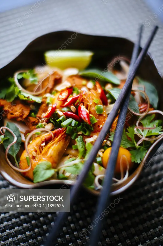 High angle close up of chopsticks on bowl with Asian food containing noodles, prawns, vegetables and chili garnish.