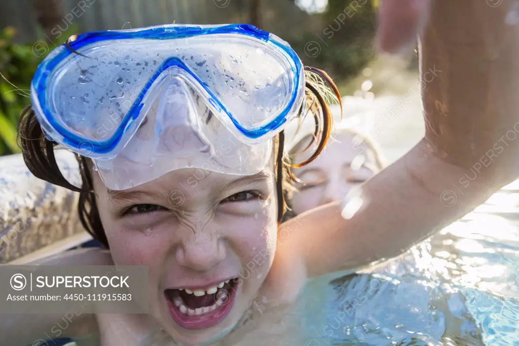 A boy in goggles laughing at the camera, in a warm swimming pool with his sister.