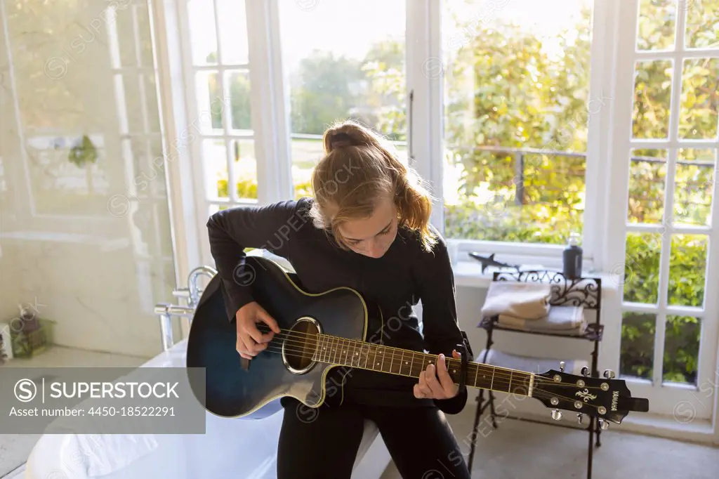 Teenage girl seated on the edge of a bathtub, playing accoustic guitar. 