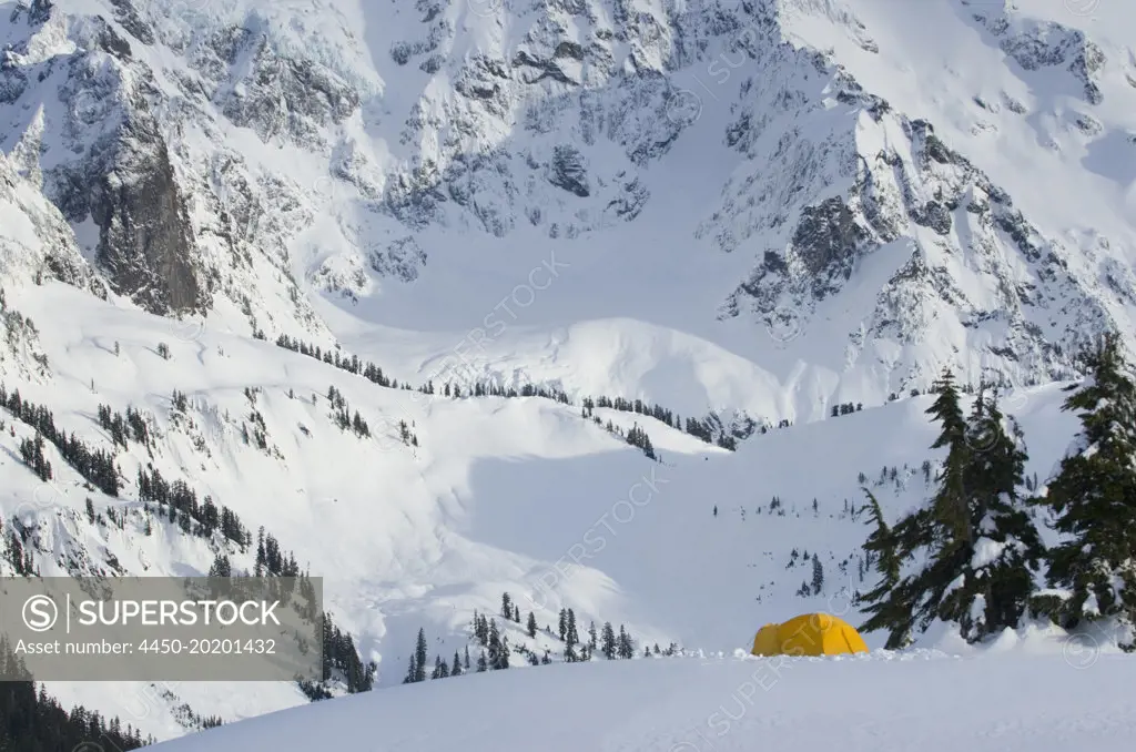 A small yellow tent pitched in deep snow on a slope, view of the steep slopes of the mountains. 